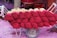Stacks of incense sticks with vibrant red dyed tops are arranged on a wooden cart. The background features various red and pink colored containers and surfaces, suggesting a setting involved in the production or sale of incense.