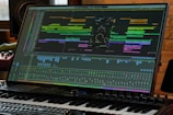 A computer monitor displays a complex digital audio workstation (DAW) interface with various tracks, levels, and audio processing tools. A piano keyboard is positioned directly in front of the monitor, indicating a music production setup. The environment appears to be a studio with professional equipment.