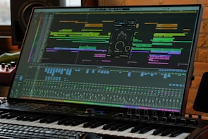 A computer monitor displays a complex digital audio workstation (DAW) interface with various tracks, levels, and audio processing tools. A piano keyboard is positioned directly in front of the monitor, indicating a music production setup. The environment appears to be a studio with professional equipment.