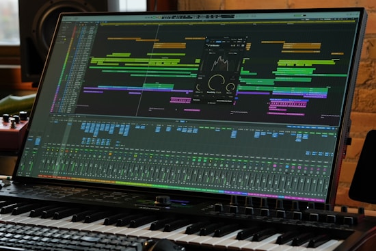 A computer monitor displays a complex digital audio workstation (DAW) interface with various tracks, levels, and audio processing tools. A piano keyboard is positioned directly in front of the monitor, indicating a music production setup. The environment appears to be a studio with professional equipment.