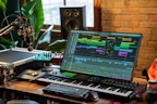 A music production setup with a large monitor displaying a digital audio workstation interface, showing multiple tracks and effects. The setup includes a keyboard, various audio equipment, a microphone, and ambient lighting. There are plants and a brick wall in the background, with natural light coming through a window.