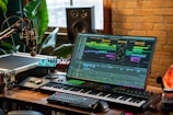 A music production setup with a large monitor displaying a digital audio workstation interface, showing multiple tracks and effects. The setup includes a keyboard, various audio equipment, a microphone, and ambient lighting. There are plants and a brick wall in the background, with natural light coming through a window.