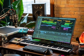 A music production setup with a large monitor displaying a digital audio workstation interface, showing multiple tracks and effects. The setup includes a keyboard, various audio equipment, a microphone, and ambient lighting. There are plants and a brick wall in the background, with natural light coming through a window.