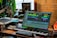 A music production setup with a large monitor displaying a digital audio workstation interface, showing multiple tracks and effects. The setup includes a keyboard, various audio equipment, a microphone, and ambient lighting. There are plants and a brick wall in the background, with natural light coming through a window.