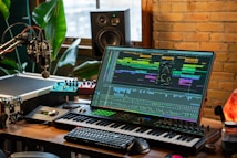 A music production setup with a large monitor displaying a digital audio workstation interface, showing multiple tracks and effects. The setup includes a keyboard, various audio equipment, a microphone, and ambient lighting. There are plants and a brick wall in the background, with natural light coming through a window.