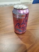 A can of La Croix sparkling water with a berry flavor rests on a wooden surface. The can is primarily pink and purple with bold branding and text indicating that it is calorie-free and naturally essenced.