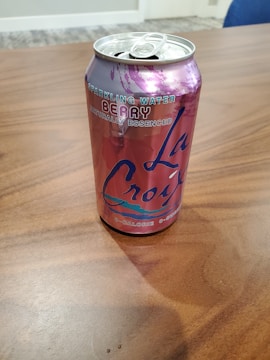 A can of La Croix sparkling water with a berry flavor rests on a wooden surface. The can is primarily pink and purple with bold branding and text indicating that it is calorie-free and naturally essenced.