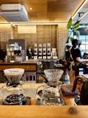 A cozy café interior with a focus on a wooden counter featuring pour-over coffee equipment. In the background, there are several coffee machines and grinders lined up against a brick and tiled wall. A person in an apron is working near the machines. The lighting is warm and inviting, with some green plant elements adding a touch of color.