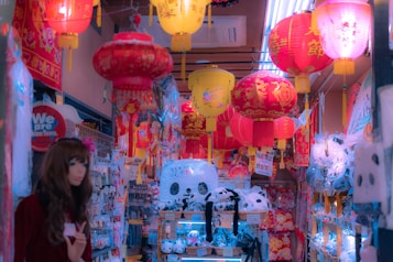 A shop filled with colorful paper lanterns in shades of red, yellow, and pink hanging from the ceiling. The shelves are stocked with various goods, including panda plush toys and other souvenir items. The atmosphere feels bustling and vibrant with intricate designs on the lanterns adding to the decorative appeal.