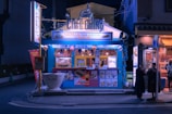 A small, vibrant street-side café with a blue exterior, offering Vietnamese egg coffee and banh mi. The café is well-lit and features colorful signage and lanterns. There are posters and menus displayed on the front. Two people stand nearby, possibly customers, under the streetlights in the evening setting.