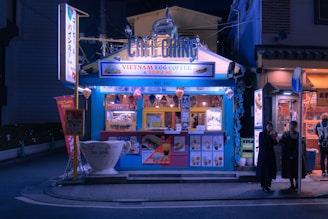 A small, vibrant street-side café with a blue exterior, offering Vietnamese egg coffee and banh mi. The café is well-lit and features colorful signage and lanterns. There are posters and menus displayed on the front. Two people stand nearby, possibly customers, under the streetlights in the evening setting.