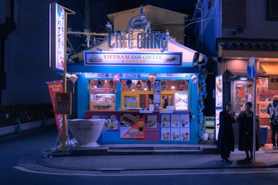 A small, vibrant street-side café with a blue exterior, offering Vietnamese egg coffee and banh mi. The café is well-lit and features colorful signage and lanterns. There are posters and menus displayed on the front. Two people stand nearby, possibly customers, under the streetlights in the evening setting.