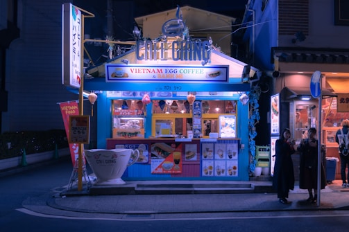 A small, vibrant street-side café with a blue exterior, offering Vietnamese egg coffee and banh mi. The café is well-lit and features colorful signage and lanterns. There are posters and menus displayed on the front. Two people stand nearby, possibly customers, under the streetlights in the evening setting.
