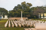A cemetery with rows of headstones arranged in neat lines, featuring a large, central stone cross. The area is surrounded by lush greenery and trees, with a backdrop of a building with a green roof.