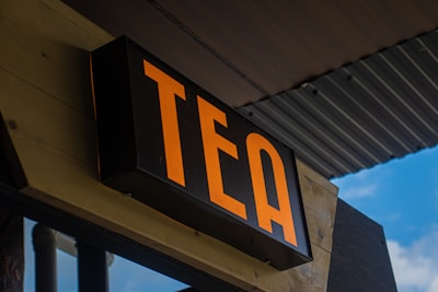 A bold black sign displaying the word 'TEA' in bright orange letters, mounted on a wooden structure. The background shows part of a corrugated metal roof and a glimpse of a blue sky.