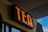A bold black sign displaying the word 'TEA' in bright orange letters, mounted on a wooden structure. The background shows part of a corrugated metal roof and a glimpse of a blue sky.