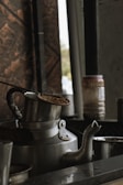 A large metal kettle with steam rising from it is placed on a countertop. The kettle appears aged with a dark handle, and there is a lid partially placed on top. In the background, there are jars and containers, while the lighting suggests an old-fashioned kitchen or tea-making setting.