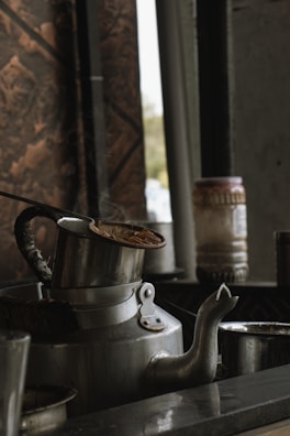 A large metal kettle with steam rising from it is placed on a countertop. The kettle appears aged with a dark handle, and there is a lid partially placed on top. In the background, there are jars and containers, while the lighting suggests an old-fashioned kitchen or tea-making setting.