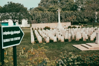 A serene cemetery with rows of white tombstones arranged in a grid pattern on a well-maintained green lawn. A prominent white memorial cross stands in the middle, surrounded by neatly trimmed bushes and colorful flowers. Trees line the background, providing a tranquil atmosphere. A sign in the foreground reads 'Madras War Cemetery' in English and another language.