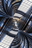 Curving architectural structures with smooth lines and large glass windows create an intricate, almost kaleidoscopic view. The perspective dramatically draws the eye upward towards the ceiling, featuring a complex grid of glass panels allowing natural light to filter through.