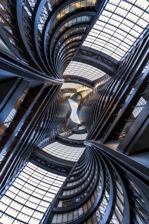 Curving architectural structures with smooth lines and large glass windows create an intricate, almost kaleidoscopic view. The perspective dramatically draws the eye upward towards the ceiling, featuring a complex grid of glass panels allowing natural light to filter through.