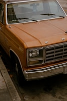 A vintage orange Ford truck is parked on the street. The truck's paint is slightly weathered, giving it a rustic feel. The front grille is prominently displayed with the emblem visible, and there is a handicap sign hanging inside on the rearview mirror. The vehicle is parked by the curb on a city street with fallen leaves scattered around.