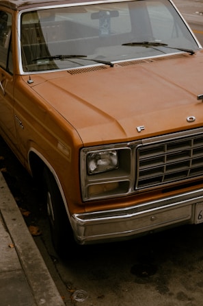 A vintage orange Ford truck is parked on the street. The truck's paint is slightly weathered, giving it a rustic feel. The front grille is prominently displayed with the emblem visible, and there is a handicap sign hanging inside on the rearview mirror. The vehicle is parked by the curb on a city street with fallen leaves scattered around.