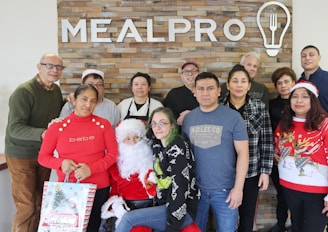 Group of people posing in front of a brick wall with a 'MEALPRO' sign. One person is dressed as Santa Claus, and others are wearing festive clothing. A woman in a red sweater holds a gift bag. The atmosphere appears to be casual and friendly.