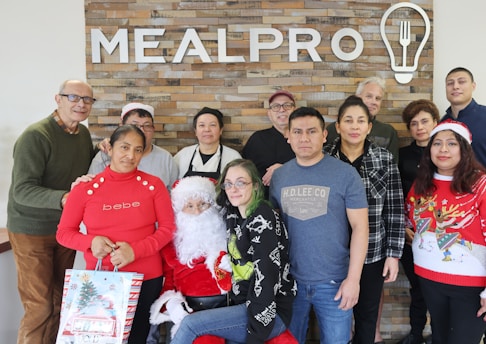 Group of people posing in front of a brick wall with a 'MEALPRO' sign. One person is dressed as Santa Claus, and others are wearing festive clothing. A woman in a red sweater holds a gift bag. The atmosphere appears to be casual and friendly.
