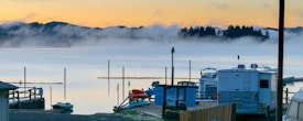 In the foreground, there are parked RVs and a few boats near a serene lake. The background features a misty lake with tall poles jutting out of the water, surrounded by silhouetted trees and rolling hills. The sky displays soft hues of orange and yellow, suggesting a sunrise or sunset.
