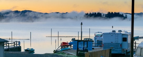 In the foreground, there are parked RVs and a few boats near a serene lake. The background features a misty lake with tall poles jutting out of the water, surrounded by silhouetted trees and rolling hills. The sky displays soft hues of orange and yellow, suggesting a sunrise or sunset.