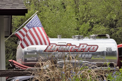 A silver industrial tanker truck with the branding 'Drain-Pro' is parked among greenery. An American flag is prominently displayed on a flagpole attached to the truck. In the foreground, there are ornamental grasses and blue flowers, while lush green trees provide a background.