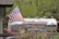 A silver industrial tanker truck with the branding 'Drain-Pro' is parked among greenery. An American flag is prominently displayed on a flagpole attached to the truck. In the foreground, there are ornamental grasses and blue flowers, while lush green trees provide a background.