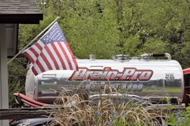 A silver industrial tanker truck with the branding 'Drain-Pro' is parked among greenery. An American flag is prominently displayed on a flagpole attached to the truck. In the foreground, there are ornamental grasses and blue flowers, while lush green trees provide a background.