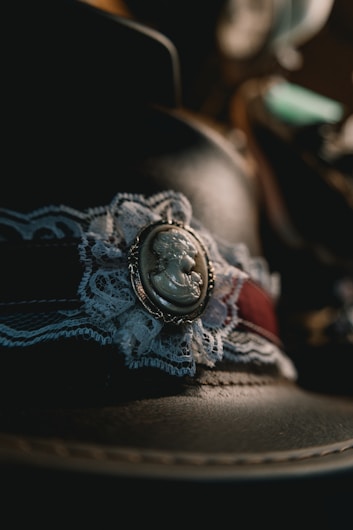A close-up of a decorative brooch set against a background of lace and fabric. The brooch features a cameo design with a profile image. The lace has intricate patterns and is paired with a band of dark reddish fabric. The lighting creates dramatic shadows and highlights.