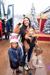 A woman is holding a baby, standing in front of a festive backdrop with artificial snow and decorated Christmas trees. Two children are beside her, one wearing a Santa hat and a winter jacket, and the other in cozy clothing. A bus and a person dressed in holiday attire are visible in the background.