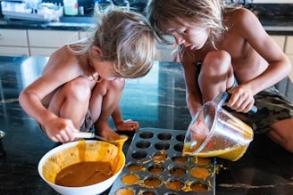 Two children with long hair are sitting on a kitchen countertop, engaging in a baking activity. One child is scooping a mixture from a white bowl while the other is pouring a liquid from a measuring jug into a muffin tray. The atmosphere is casual and playful.