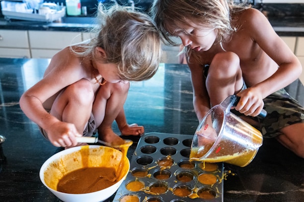 Two children with long hair are sitting on a kitchen countertop, engaging in a baking activity. One child is scooping a mixture from a white bowl while the other is pouring a liquid from a measuring jug into a muffin tray. The atmosphere is casual and playful.