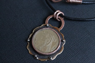 A pendant featuring a coin with an engraved profile of a figure, surrounded by a metal frame and attached to a black cord necklace. The metal frame has a scalloped edge and metallic rings.