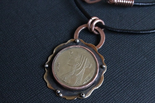 A pendant featuring a coin with an engraved profile of a figure, surrounded by a metal frame and attached to a black cord necklace. The metal frame has a scalloped edge and metallic rings.