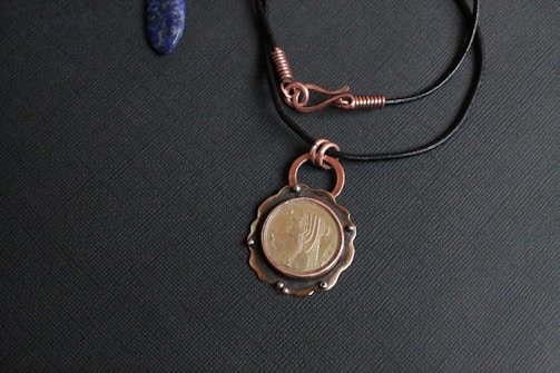 A necklace with a pendant featuring a coin design with a profile of a woman. The pendant is attached to a black cord with copper wire accents. The surface appears textured and the overall color scheme is dark with metallic highlights.