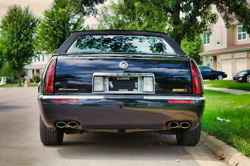 A black luxury sedan parked on a suburban street, featuring an Illinois license plate. The car has dual exhausts and the badge 'ESC' on the back. The background includes green trees and houses with well-manicured lawns.