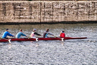 A team of five rowers are paddling a long, narrow boat on a body of water. The rowers are dressed in sporty attire, and the boat is red. In the background, a textured, stone wall runs parallel to the water's surface.