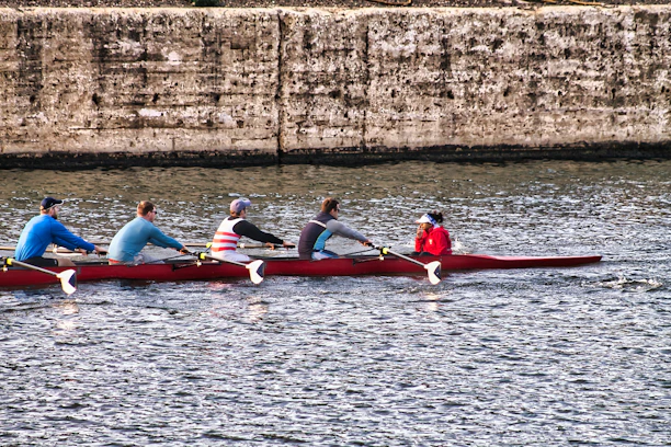 A team of five rowers are paddling a long, narrow boat on a body of water. The rowers are dressed in sporty attire, and the boat is red. In the background, a textured, stone wall runs parallel to the water's surface.