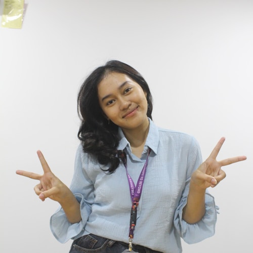 A person with long, dark hair, wearing a light blue shirt and jeans, poses against a plain white background. They are smiling and gesturing with both hands, making a peace sign with their fingers. A lanyard with an ID card is hanging around their neck.