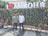 Two men are standing in front of a wall covered with plants. Above the wall, there are large letters that read 'I Love (with a heart symbol) AMROHA'. They are posing for the photograph in a bright, outdoor setting. There is another person seated on a bench to the left, near potted plants.