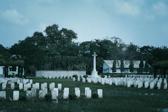 A cemetery with numerous white headstones neatly arranged in rows. In the center stands a prominent cross monument. The background features dense green trees and a white building with a roof. The overall scene is calm and somber, with overcast skies adding to the atmosphere.
