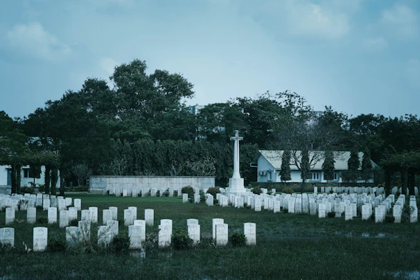 A cemetery with numerous white headstones neatly arranged in rows. In the center stands a prominent cross monument. The background features dense green trees and a white building with a roof. The overall scene is calm and somber, with overcast skies adding to the atmosphere.