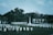 A cemetery with numerous white headstones neatly arranged in rows. In the center stands a prominent cross monument. The background features dense green trees and a white building with a roof. The overall scene is calm and somber, with overcast skies adding to the atmosphere.
