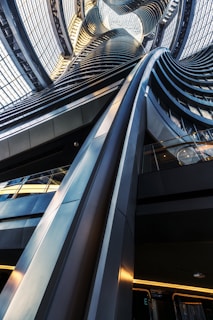 A striking architectural interior featuring twisting, undulating metal and glass structures that extend upward, capturing light and reflections. The design is modern and futuristic, showcasing intricate shapes and engineering.